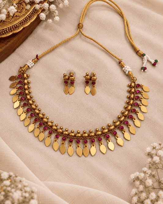 Antique gold look Elakkathali necklace set featuring pink stones, hanging leaf-shaped motifs and matching earrings arranged in a flatlay
