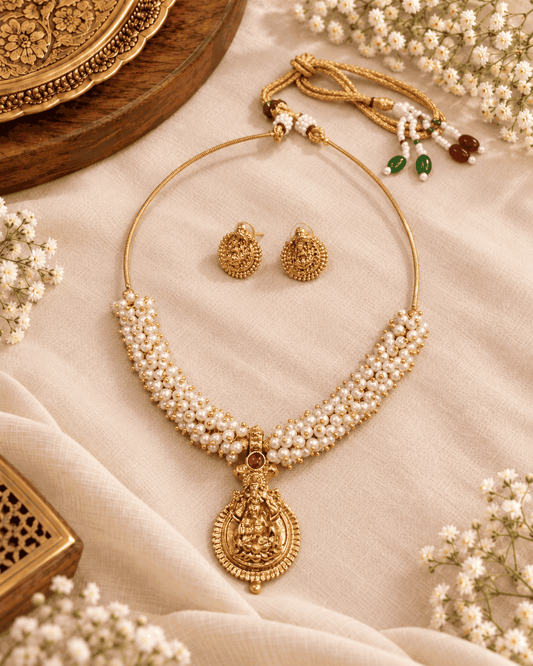 Gold look Lakshmi temple necklace with multiple rows of white pearl-like beads, pink stone pendant and matching earrings arranged on fabric
